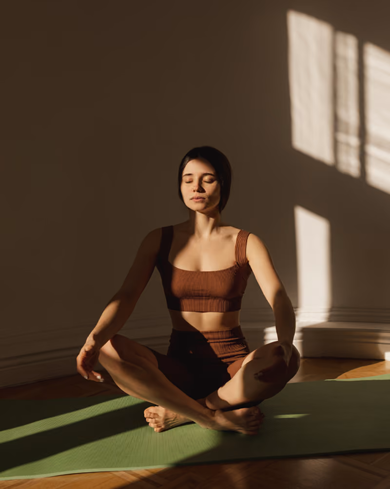 Eine Frau in Sportkleidung meditiert auf einer grünen Yogamatte im Sonnenschein.