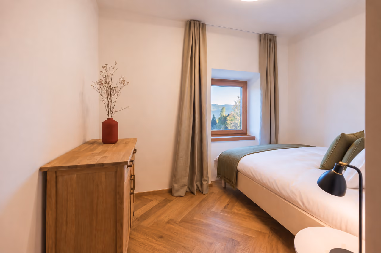 Schlafzimmer im Gregorhof mit Holzkommode, Leselampe und Fenster mit Blick in den Wald.