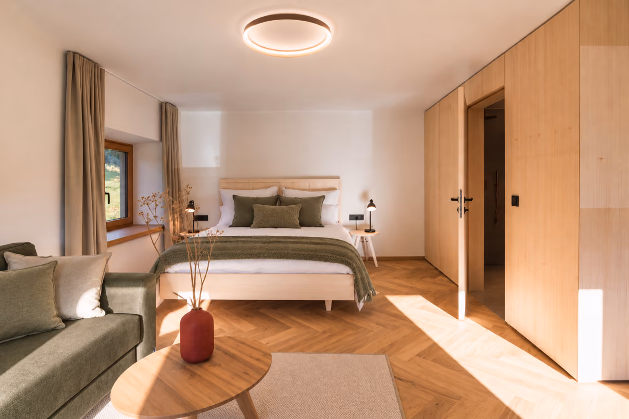 Helles Schlafzimmer im Gregorhof mit Doppelbett, Sofa und kleinem Holztisch auf Fischgrätparkett.