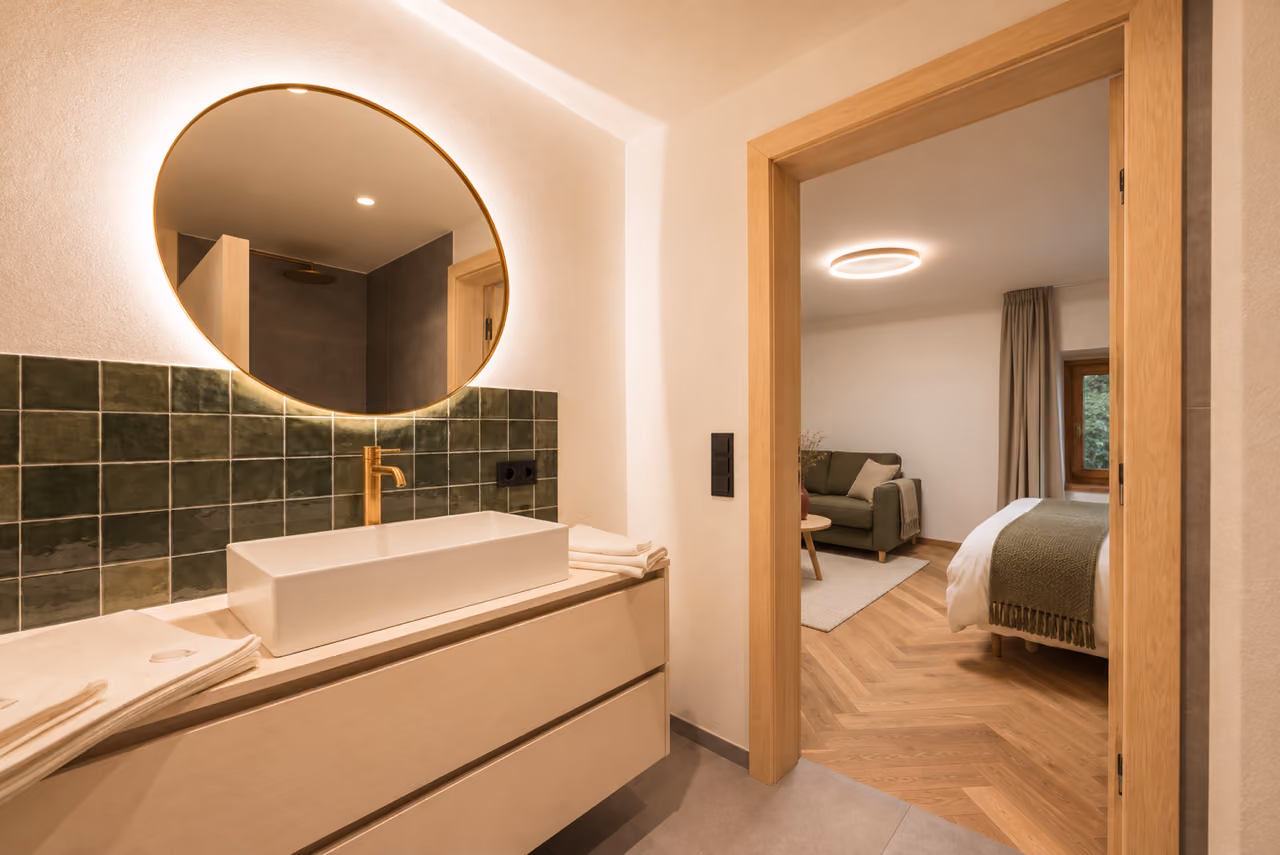 Badezimmer im Gregorhof mit rundem beleuchtetem Spiegel, grünen Fliesen und Blick ins Schlafzimmer.