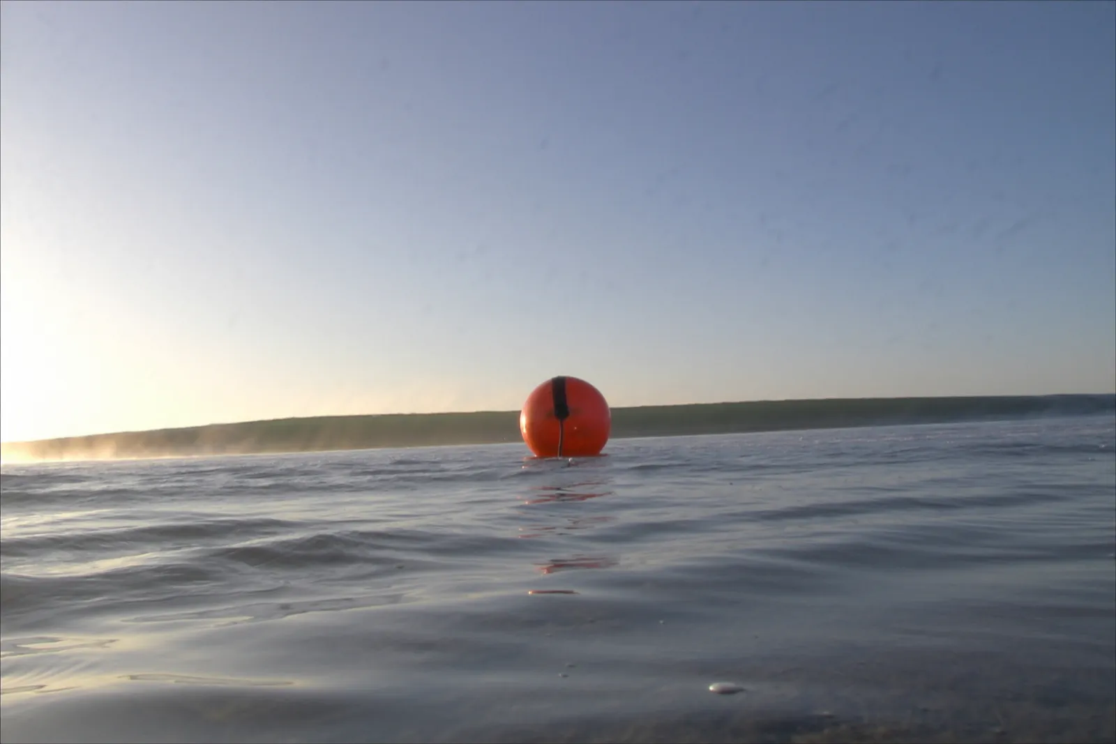 Open sea in daylight, grey-blue swell to a level horizon, one orange buoy or work boat on the water off the coast of