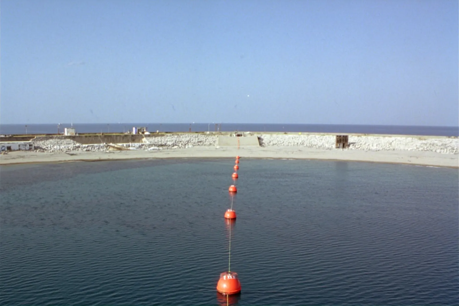 Mediterranean coast at a subsea cable landing in daylight, one orange marker or structure by calm blue sea near Genoa