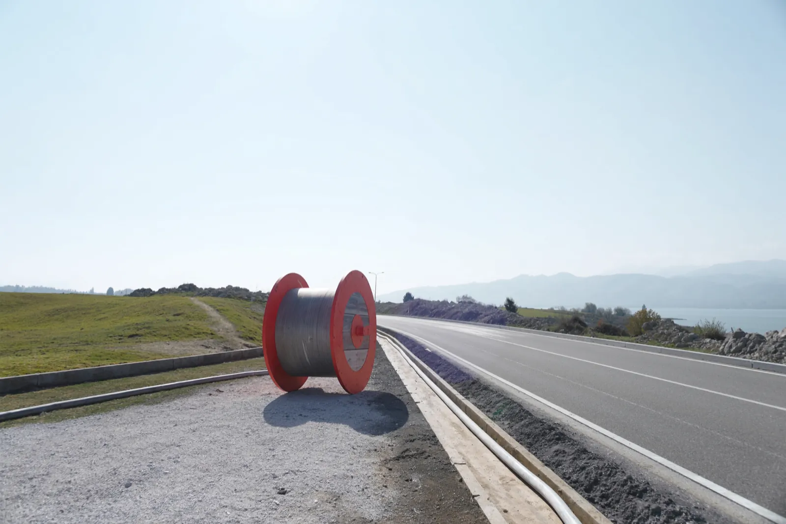 Road through a valley in daylight, cable works on the verge with orange markers and equipment between Genoa and Milan