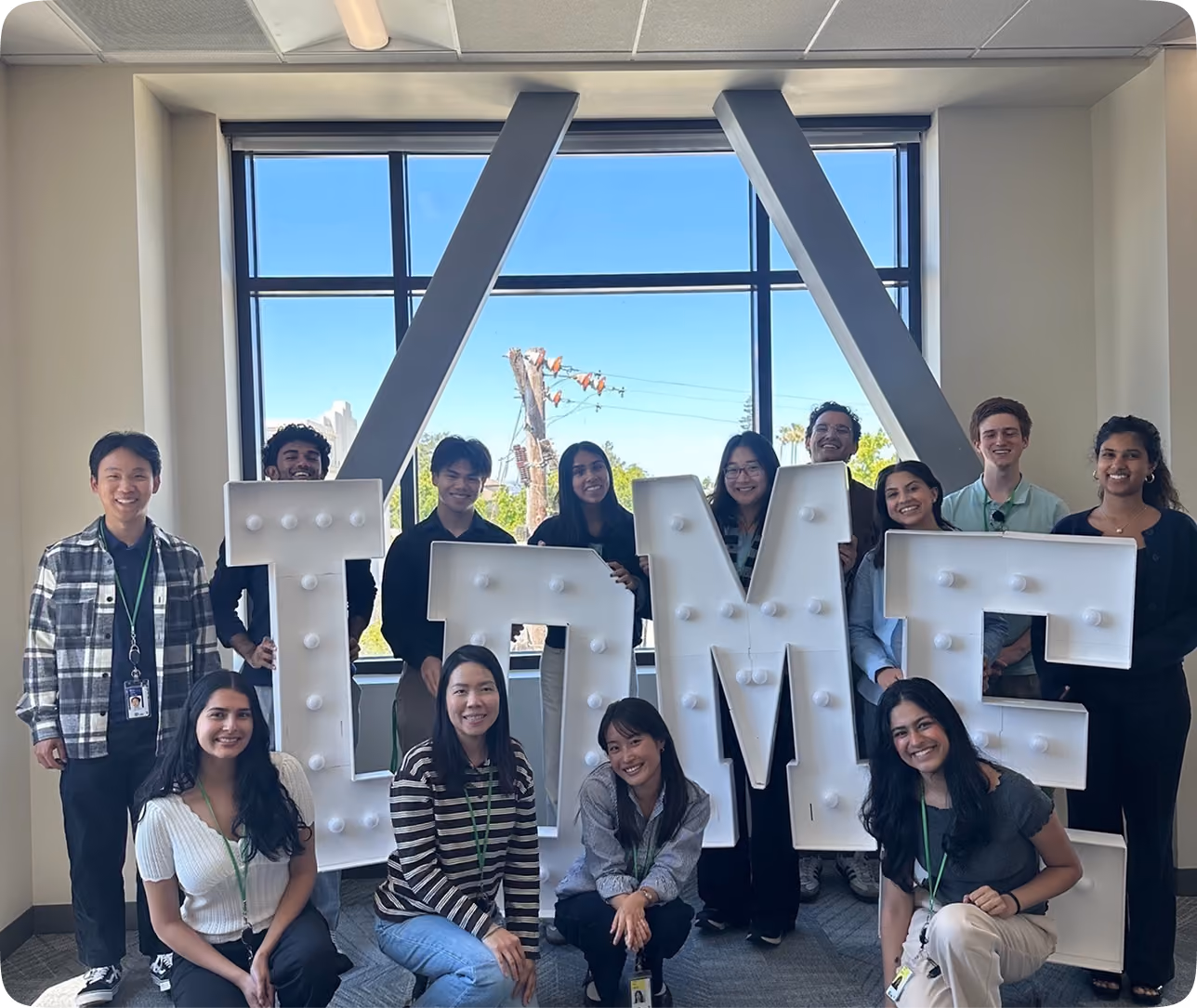 Group of thirteen smiling young adults posing indoors around large illuminated letters spelling 'LAME' with a large window and blue sky in the background.