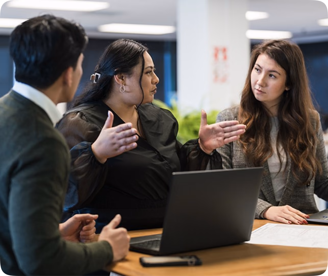Three colleagues engaged in a discussion around a table with an open laptop in an office setting, with one woman gesturing to emphasize her point.
