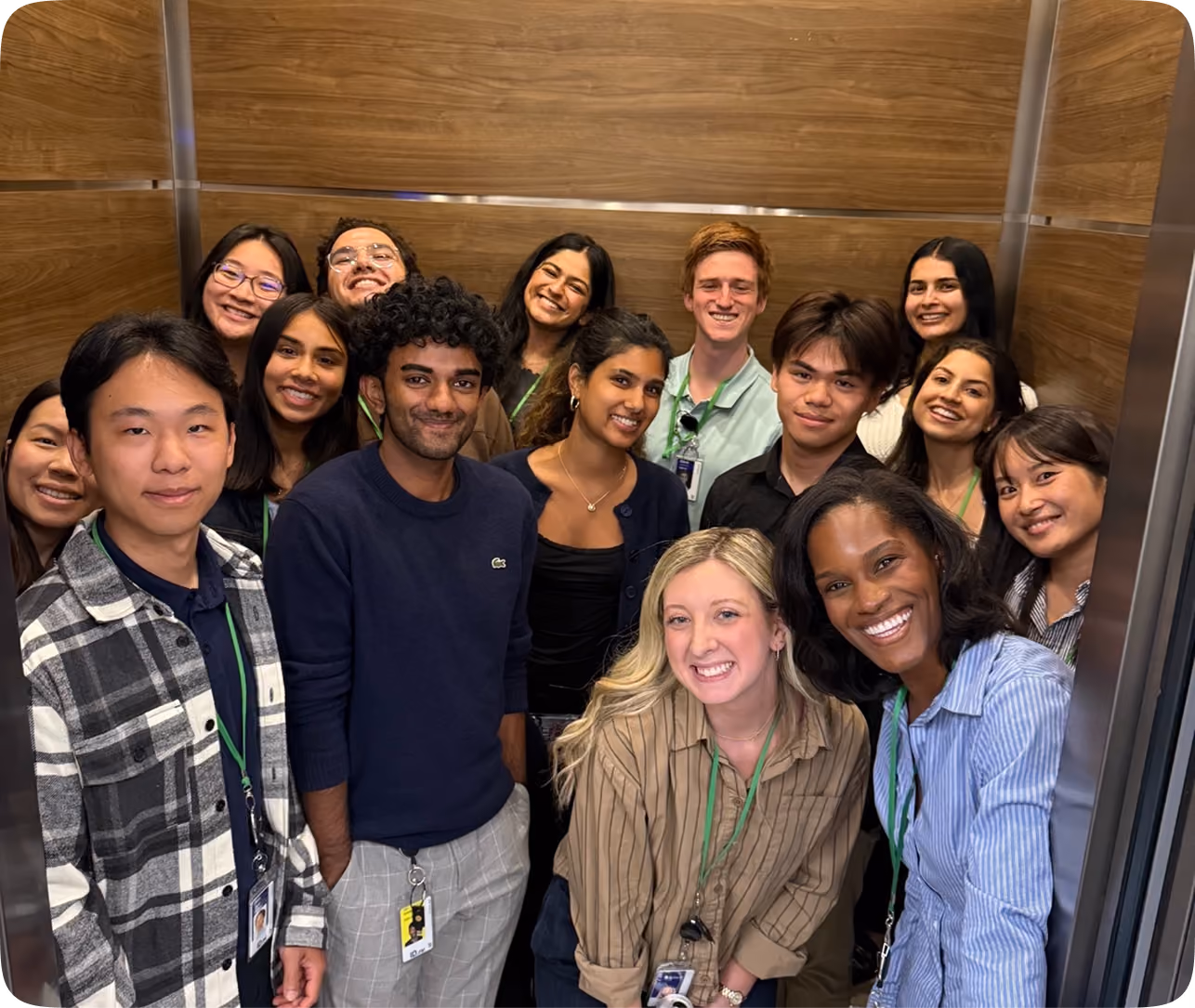 A diverse group of 14 young adults smiling closely together inside a small elevator with wood-paneled walls, wearing casual clothes and ID badges.