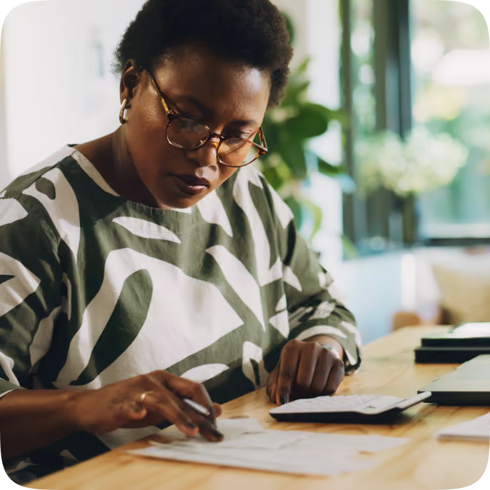 A focused woman wearing glasses and a patterned shirt uses a calculator while reviewing papers at a wooden desk in a bright room with plants and a window in the background.
