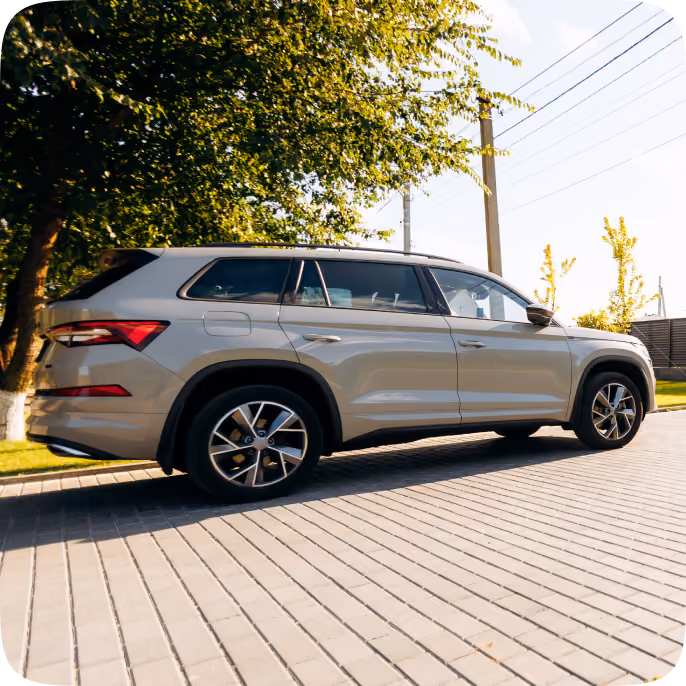 Side view of a beige SUV parked on a paved area next to a tree, with power lines and a fence in the background under a clear sky.