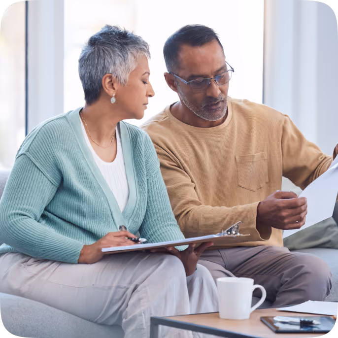 A middle-aged woman with short gray hair and a man with glasses sit on a couch reviewing documents together, with a clipboard and a cup on the table in front of them.