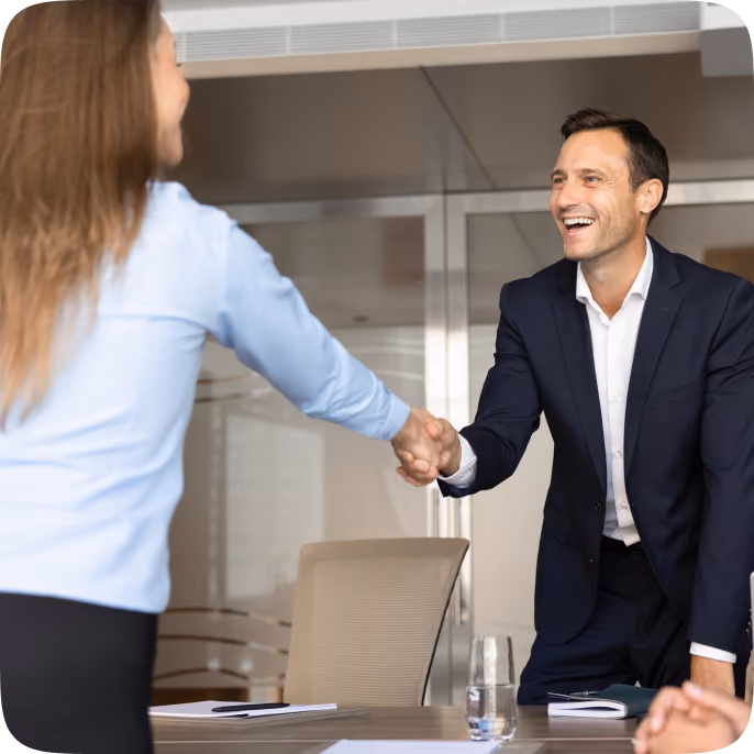 A man in a dark suit smiling and shaking hands with a woman in a light blue shirt in a modern office setting.
