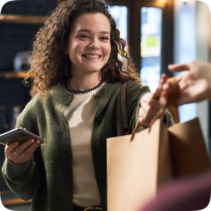 Smiling young woman with curly hair holding a smartphone in one hand and reaching out to take a brown shopping bag handed to her.