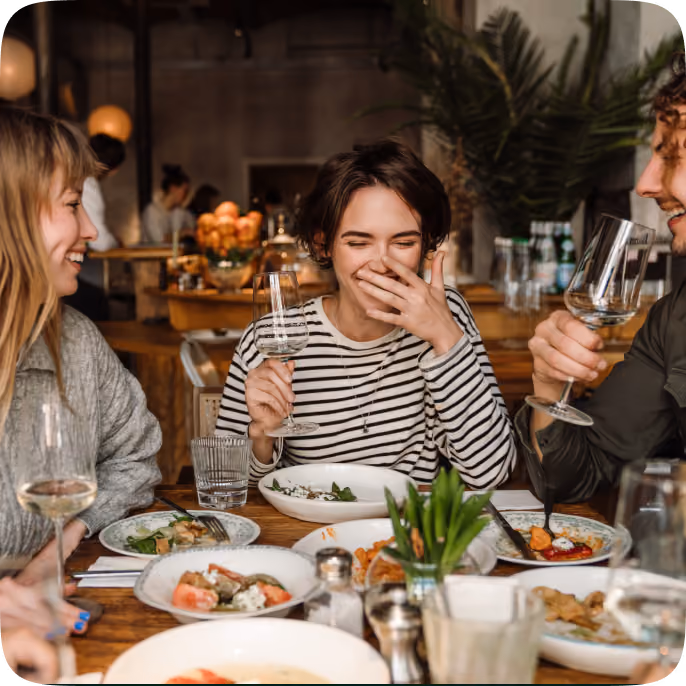 Three friends sitting at a wooden table in a restaurant, enjoying food and wine while laughing and smiling together.