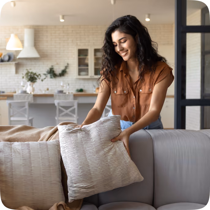 A smiling woman with long dark hair arranging a textured beige pillow on a light gray sofa in a modern living room with a kitchen and dining area in the background.