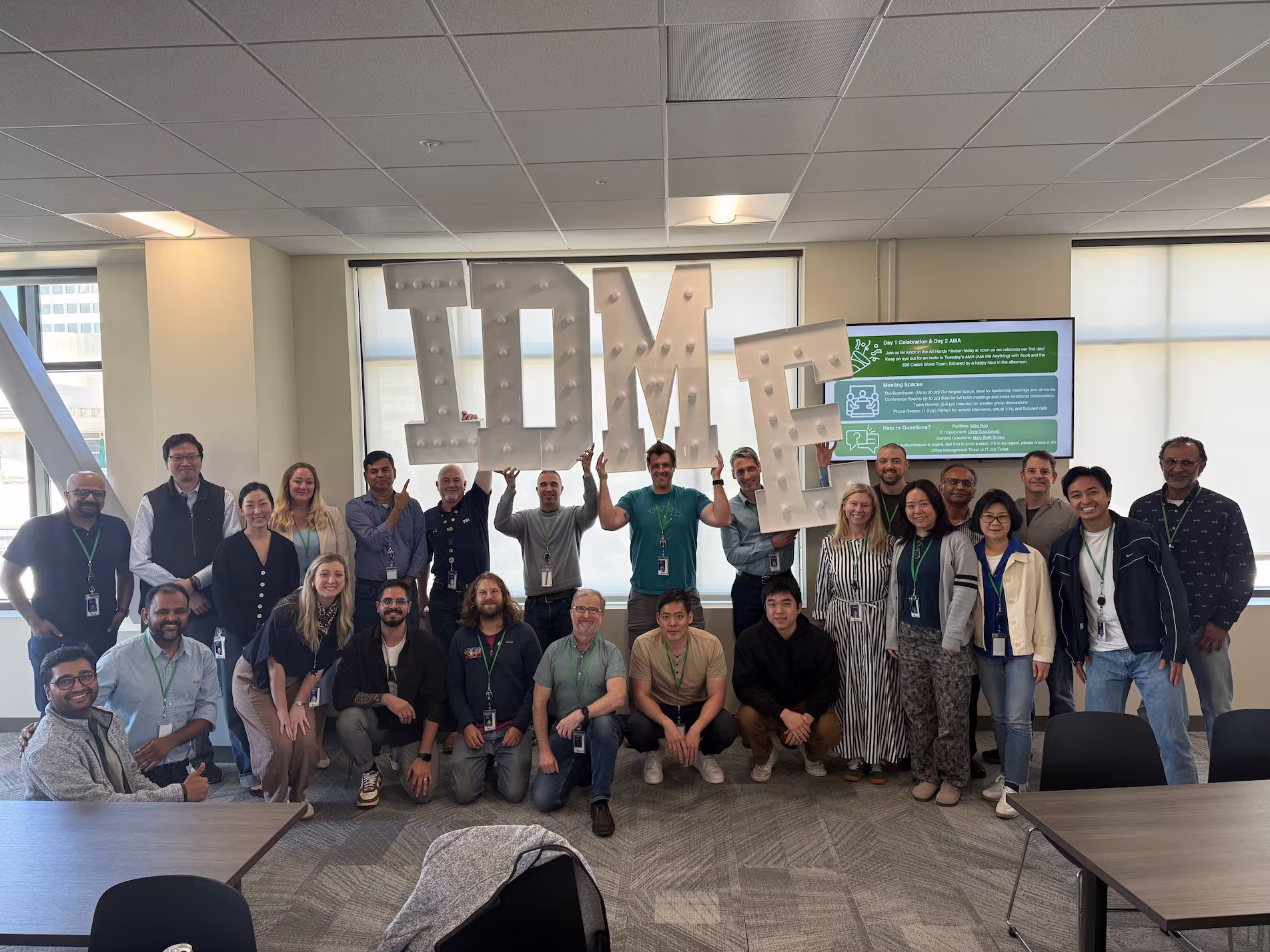 A diverse group of 26 people in a bright office posing and smiling for a group photo. Several individuals in the back row hold large illuminated letters spelling 'IDME'.