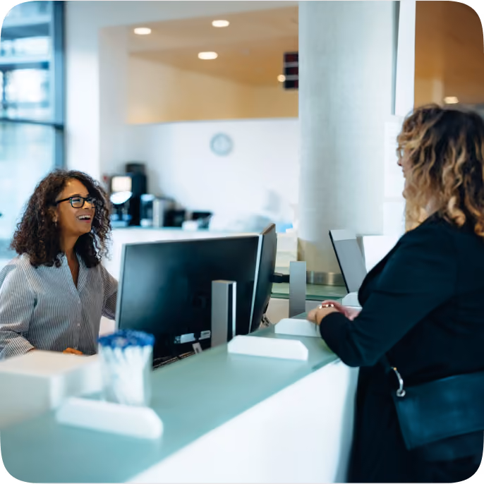 A receptionist woman with curly hair and glasses smiling and talking to a woman visitor standing at a modern, light-colored reception desk with computer monitors in an office lobby.