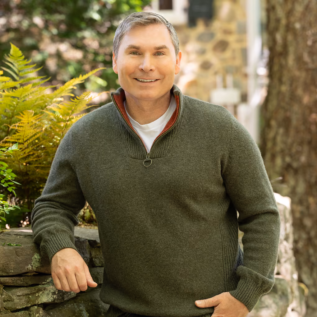 Middle-aged man with short gray hair wearing a green quarter-zip sweater over a white shirt, smiling and leaning on a stone wall outdoors with green ferns and trees in the background.