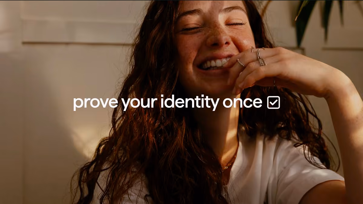 Smiling woman with long curly hair in white shirt, resting her hand near her face with a relaxed expression, with text overlay saying 'prove your identity once' and a checkmark icon.