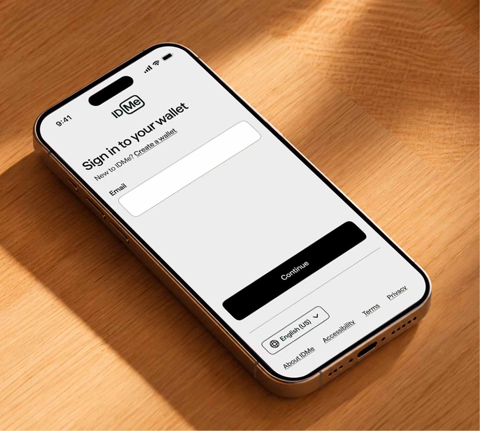 Smartphone on a wooden surface displaying the ID.me sign-in screen with fields for email entry, a black continue button, language selection dropdown set to English (US), and links for About ID.me, Accessibility, Terms, and Privacy.