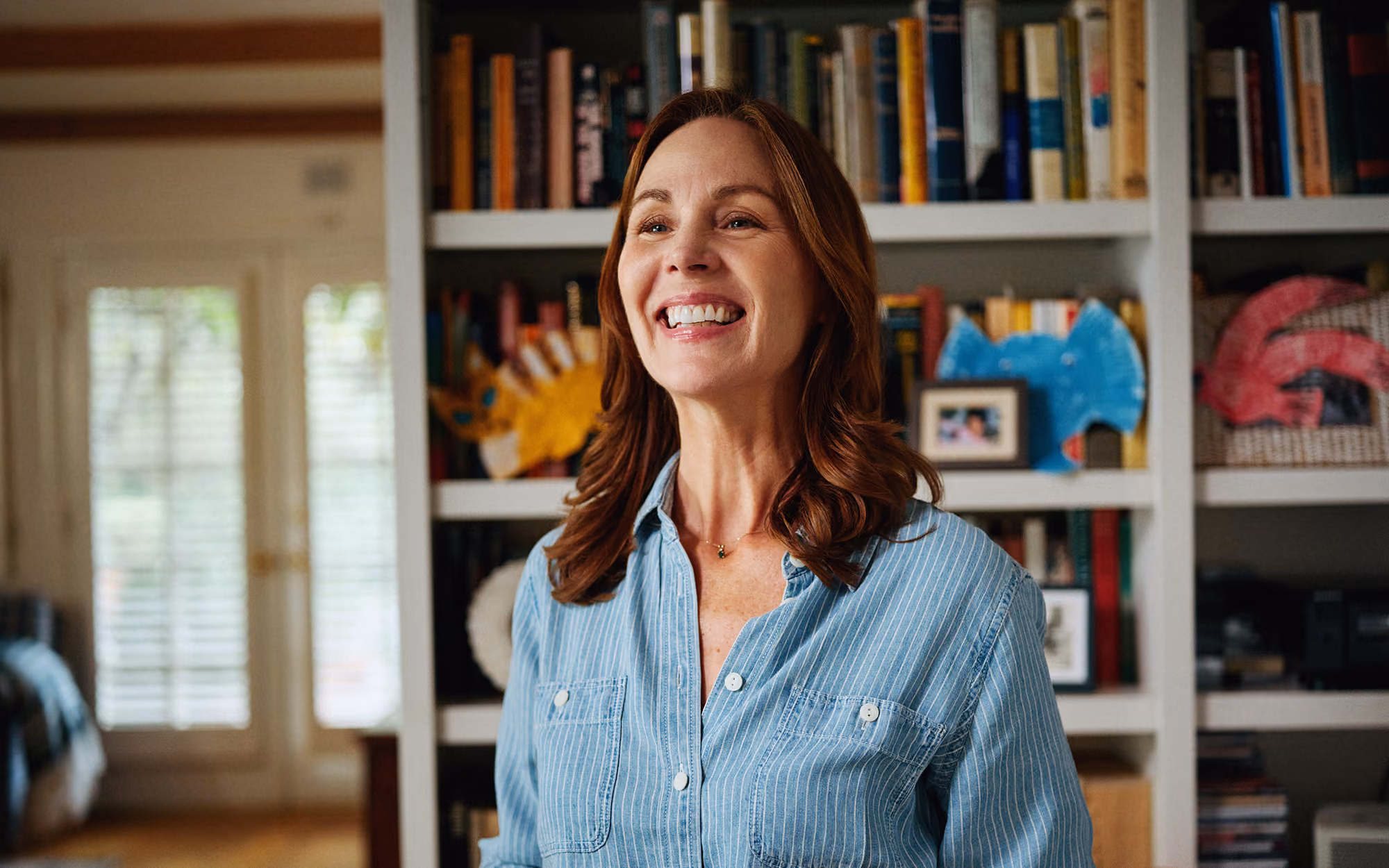 Smiling woman with reddish-brown hair in a blue striped shirt standing indoors in front of shelves with books and colorful hand-shaped art.