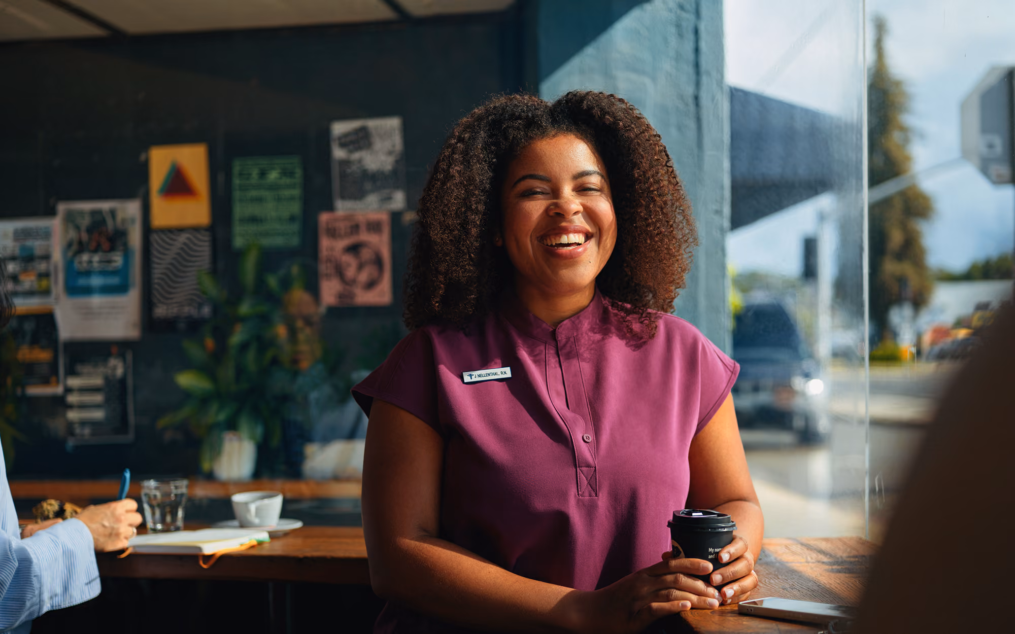 Smiling woman with curly hair in a purple blouse holding a coffee cup, sitting by a window in a cozy café.