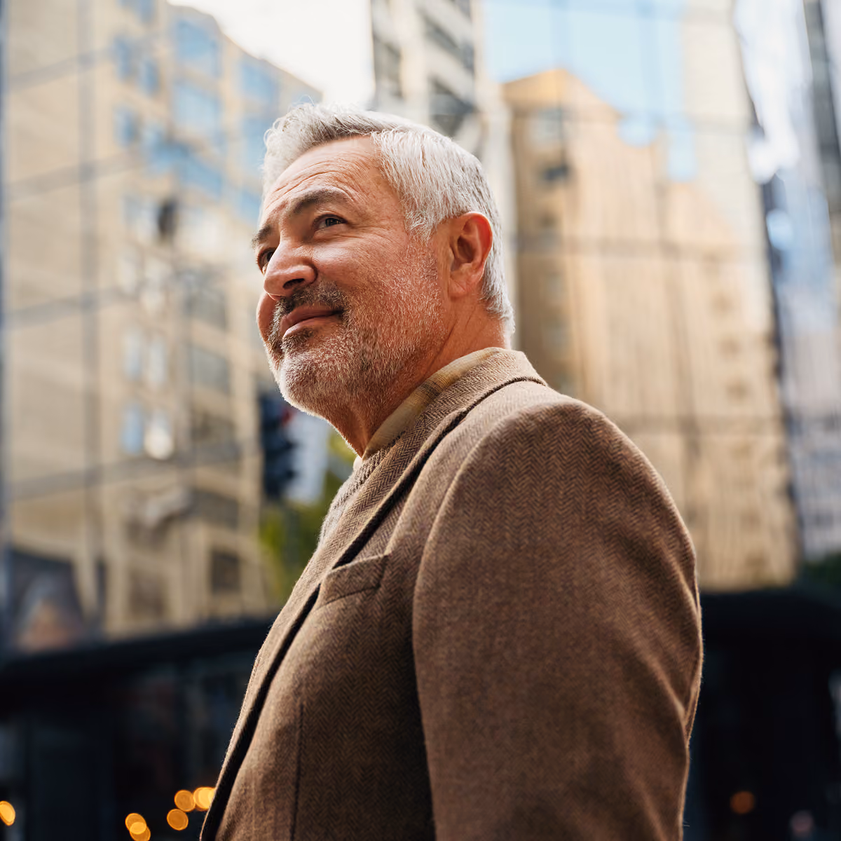 Older man with gray hair and beard wearing a brown tweed jacket, looking thoughtfully upward against a blurred urban backdrop of tall buildings and city lights.