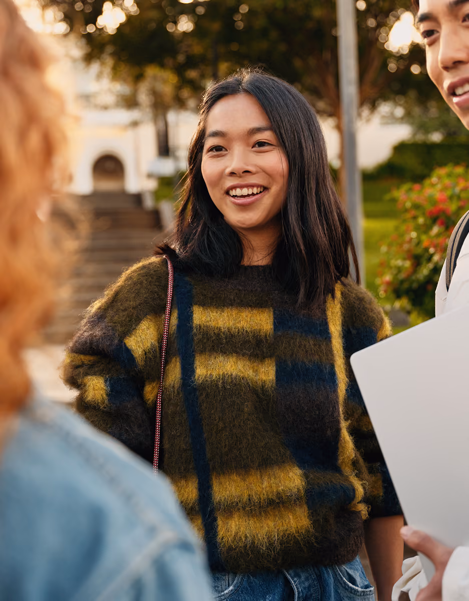 Three young adults standing outdoors in a park or campus setting, talking and smiling. One woman with long dark hair wears a fuzzy plaid sweater and jeans, another person with curly blonde hair is seen from behind, and a man holding a laptop is partially visible on the right.