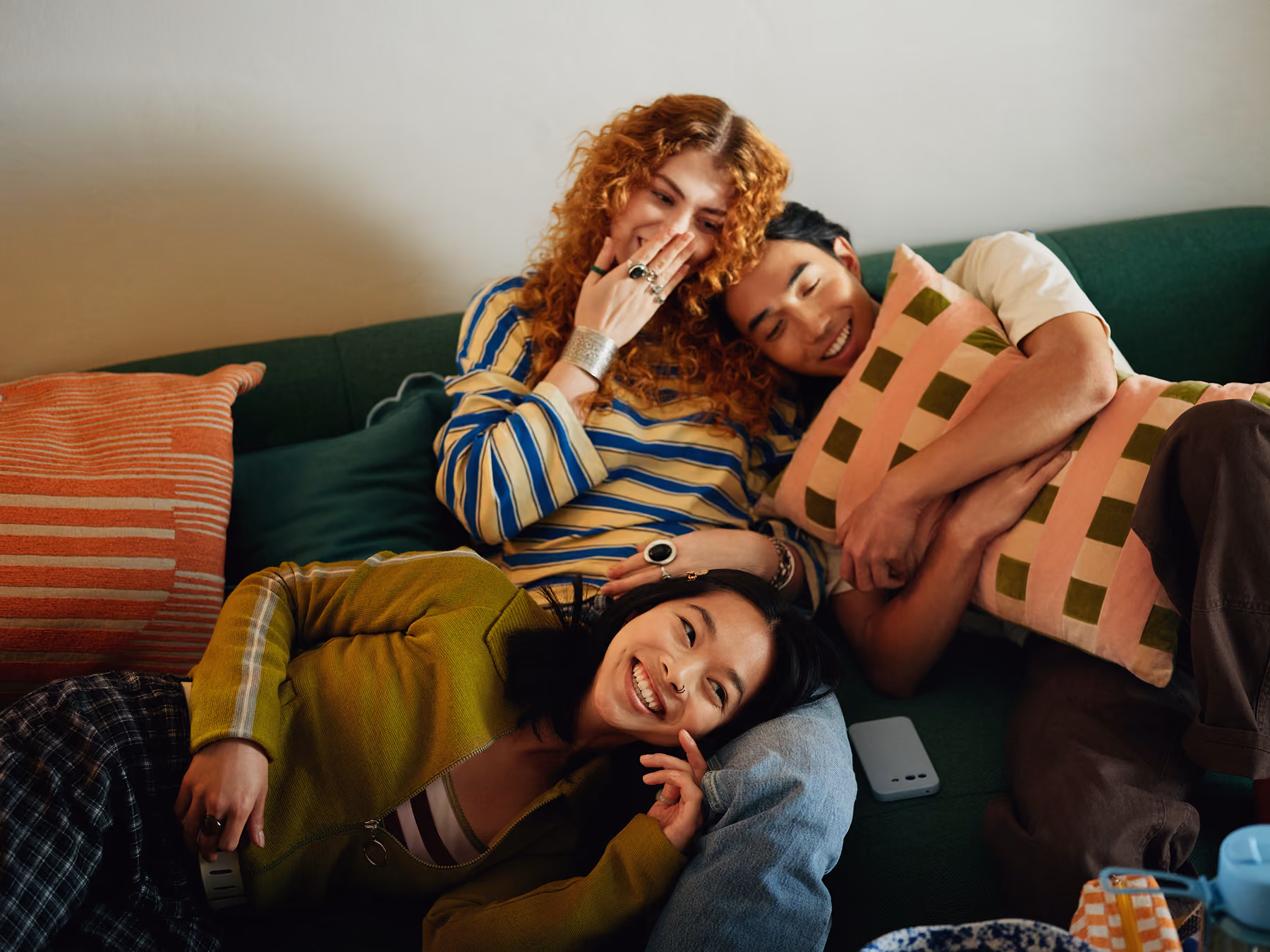 Three friends relaxing closely on a green couch indoors: a curly red-haired woman in a striped shirt laughs with her hand near her face, a man hugs a checkered cushion while smiling with his eyes closed, and a woman wearing a green sweater lays her head on the man's lap smiling.