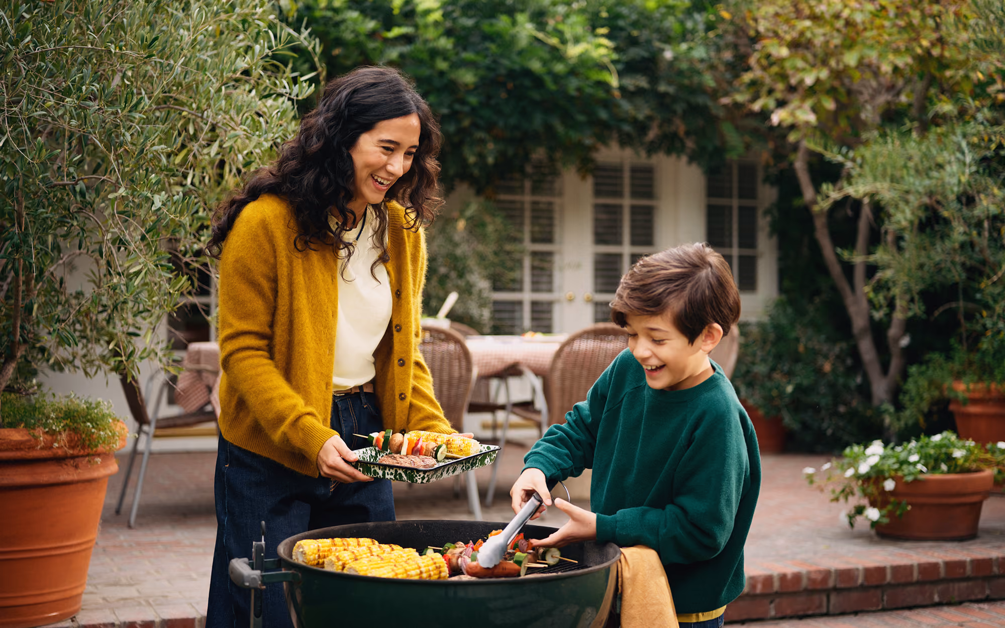 A smiling woman in a yellow cardigan holds a plate of grilled vegetables and corn while a young boy in a green sweatshirt uses tongs to cook skewers and corn on a round outdoor grill in a backyard patio surrounded by plants.