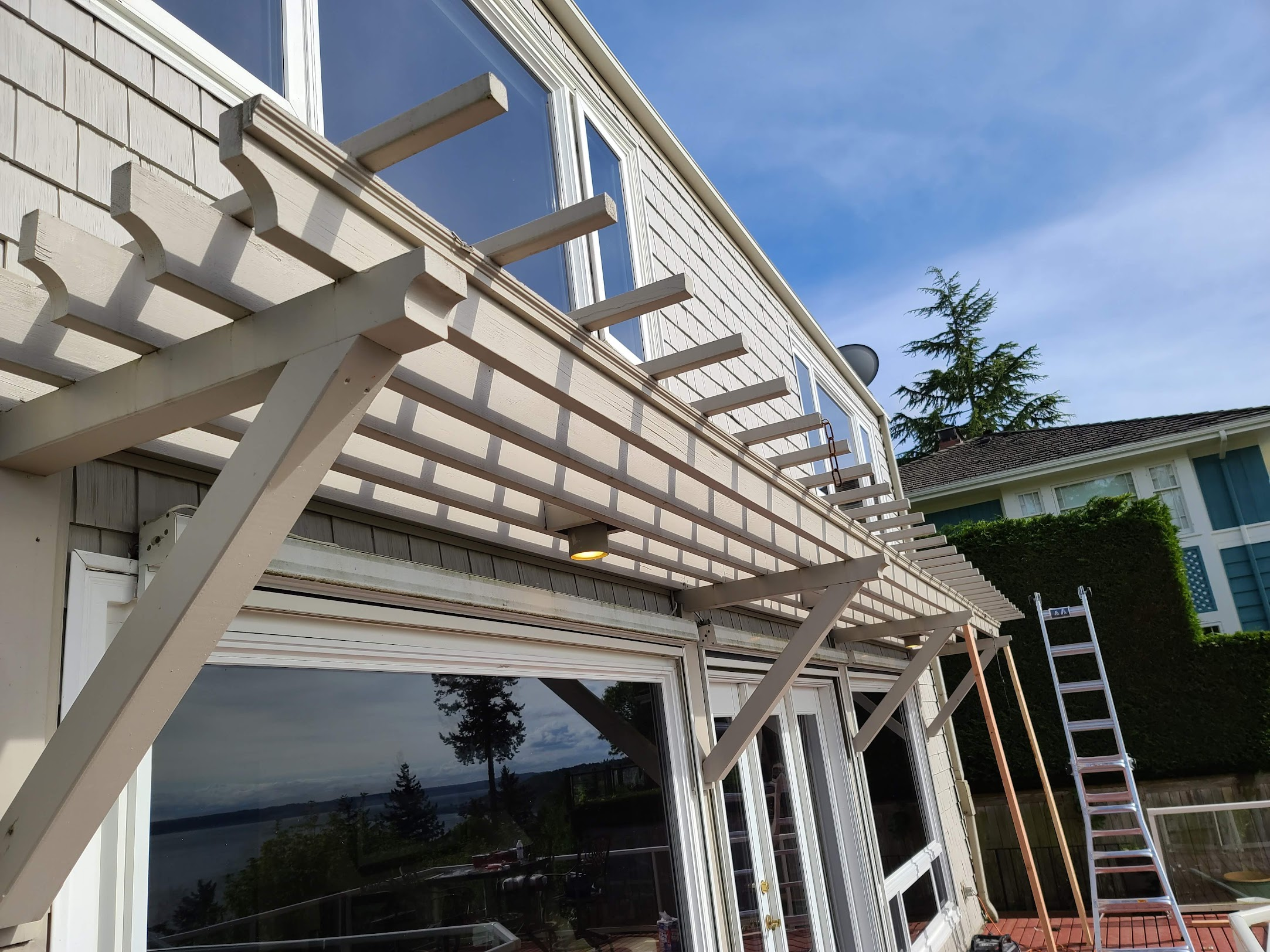 Wooden trellis structure undergoing repair along the side of a home above large windows.