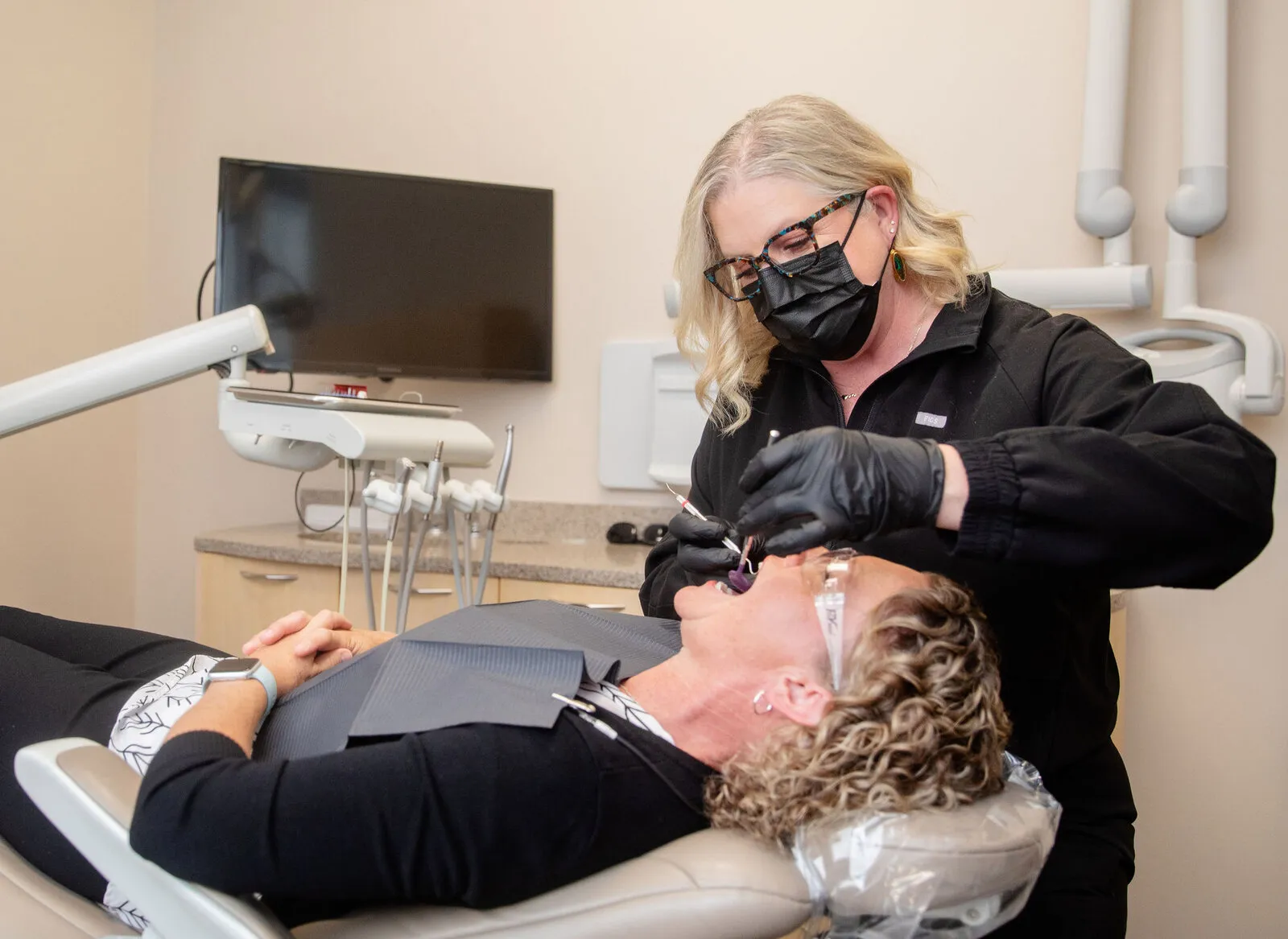 Dental hygienist in black scrubs and mask performing a cleaning on a patient in a modern dental office