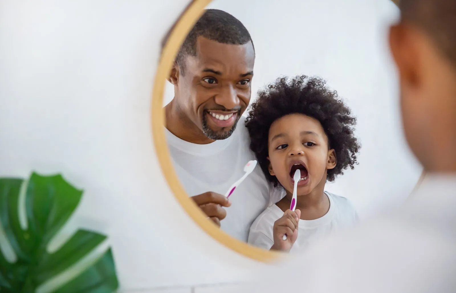 Father and young son brushing their teeth together, reflected in a round bathroom mirror
