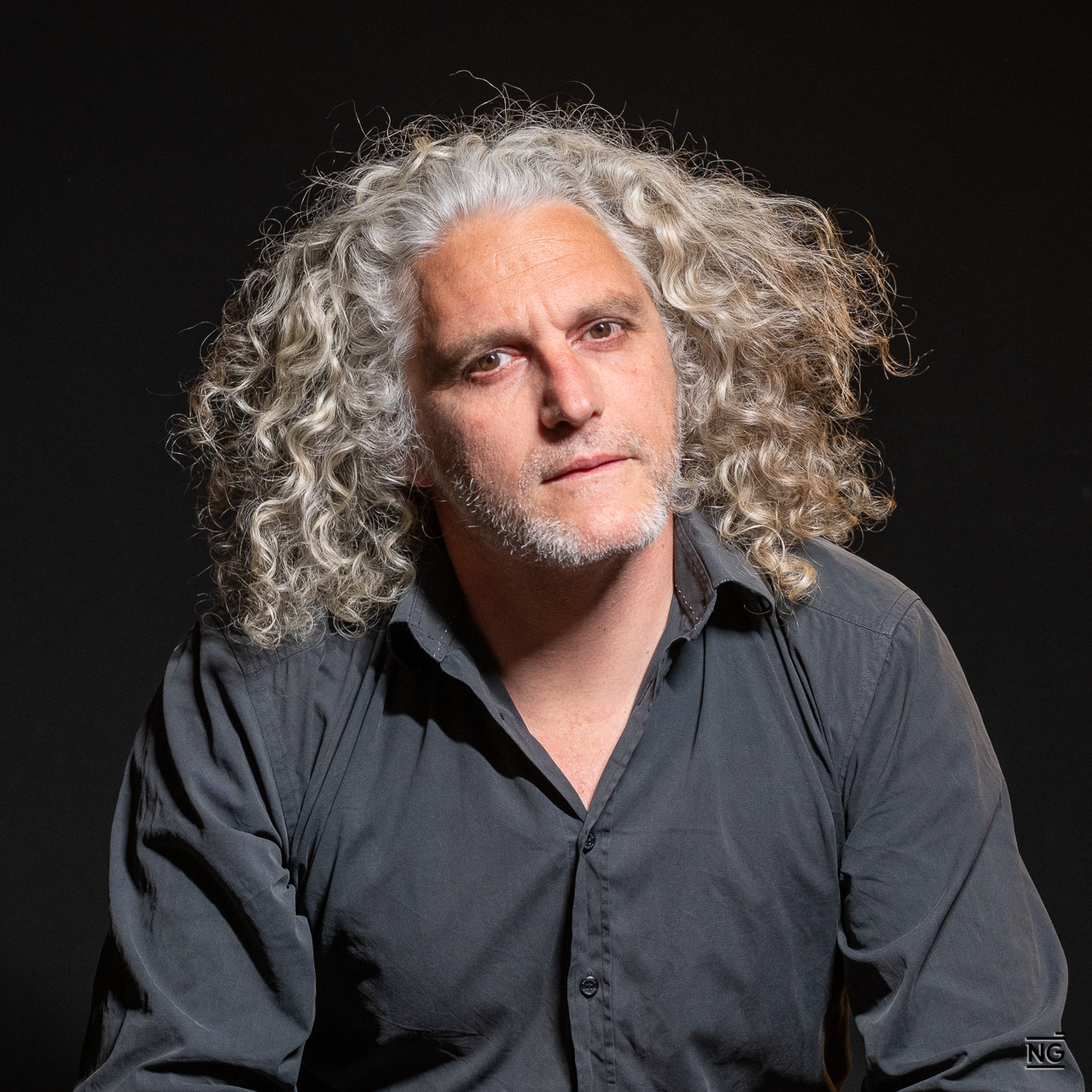 Homme avec cheveux longs et bouclés gris et barbe naissante, portant une chemise noire, sur fond noir.