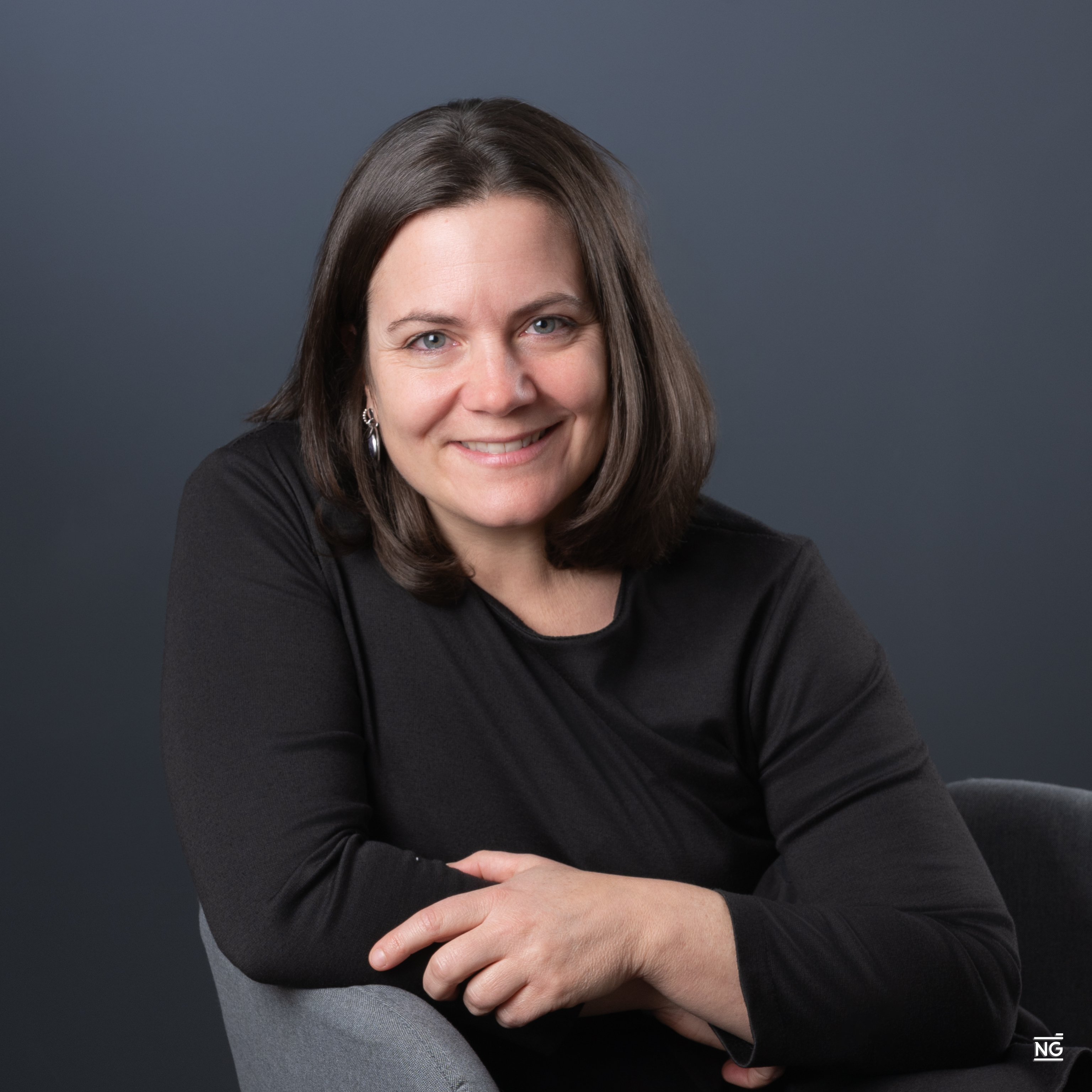 Portrait d'une femme souriante aux cheveux bruns mi-longs, portant un haut noir, assise sur une chaise grise avec un fond gris uni.
