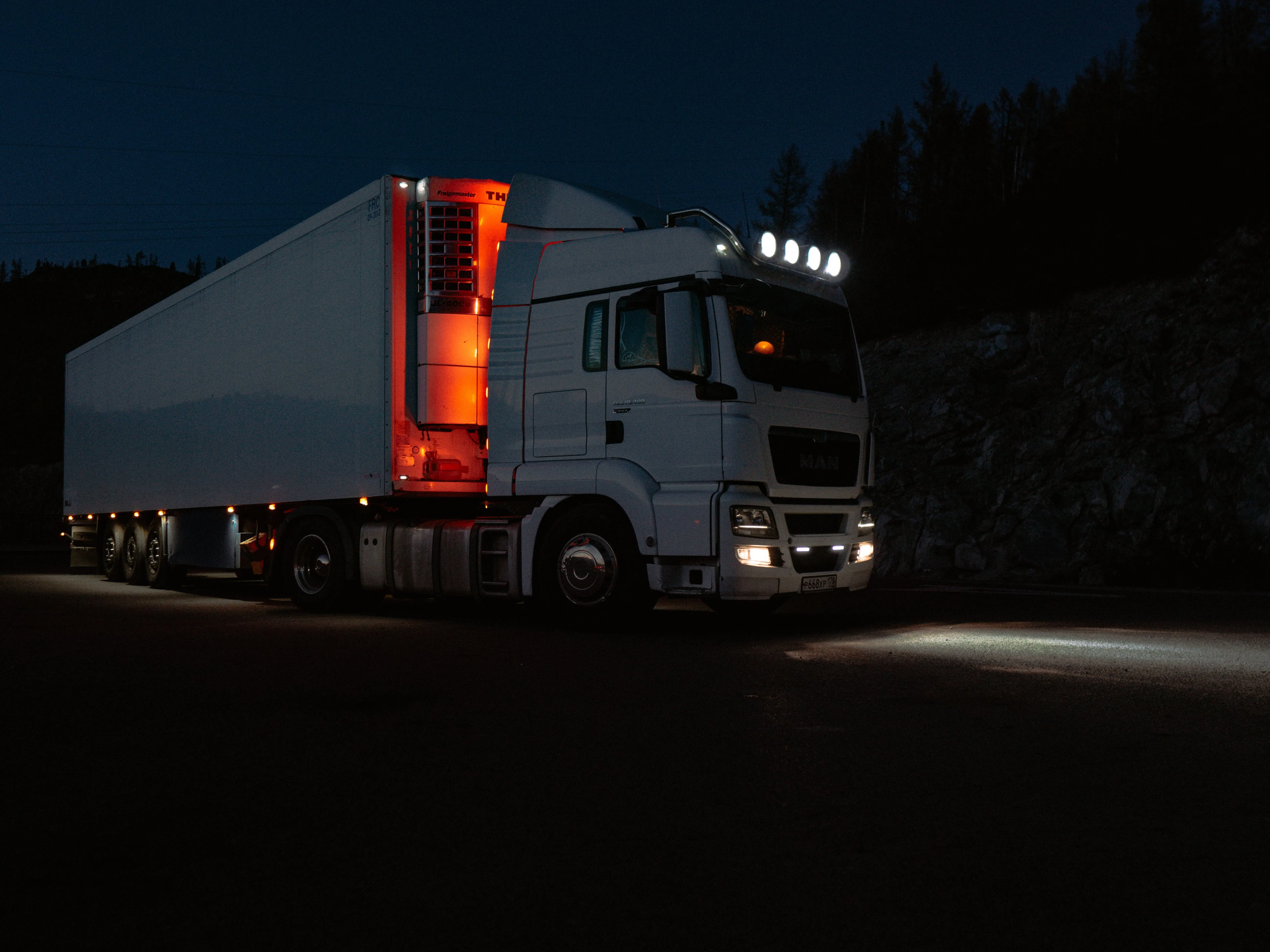White semi-truck with trailer illuminated by front headlights and red interior trailer lights parked at night on a road near rocky terrain and trees.