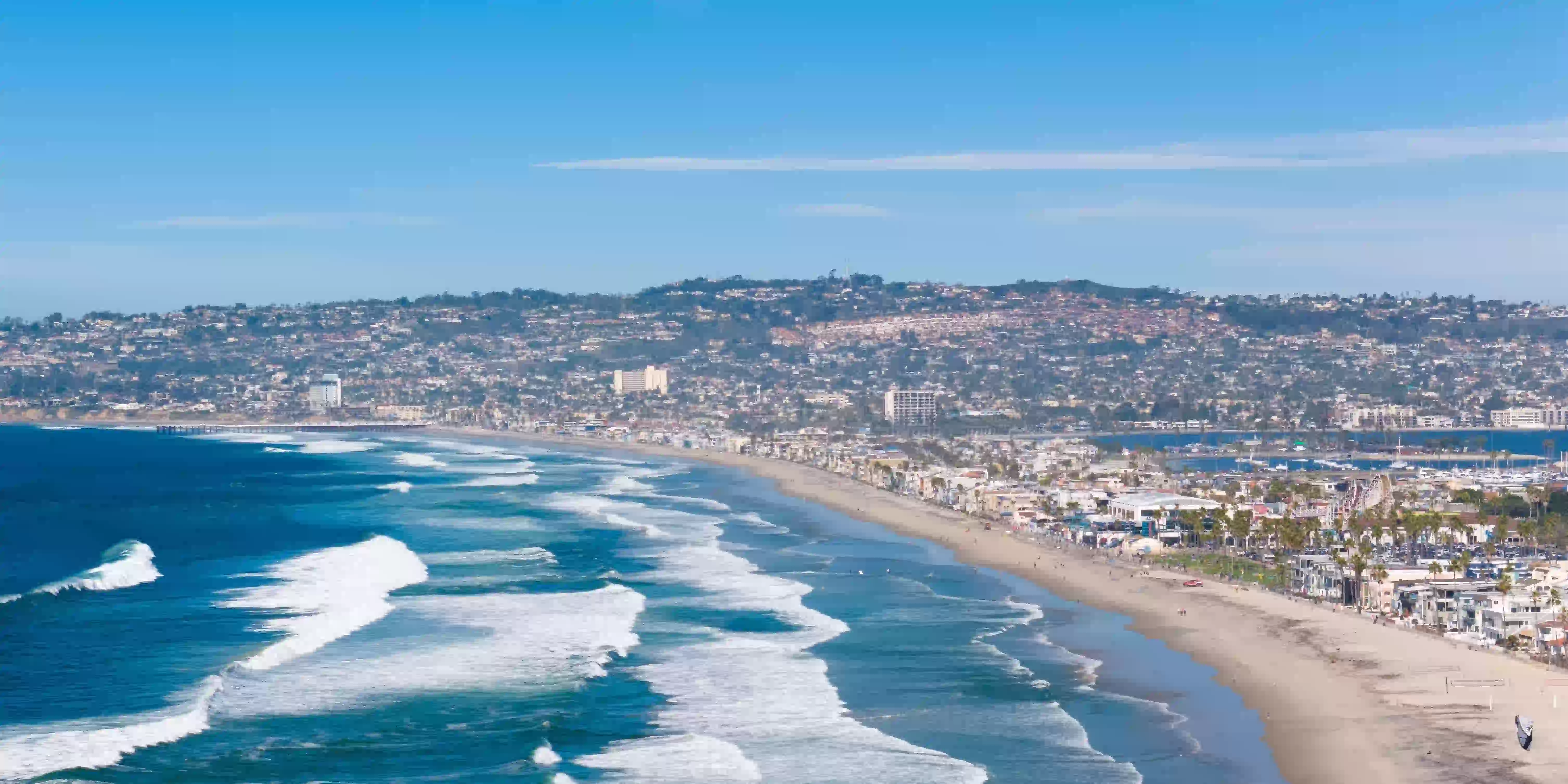 A wide sandy beach with large waves crashing onto shore under a clear blue sky, with a cityscape and hills in the background along the coastline.