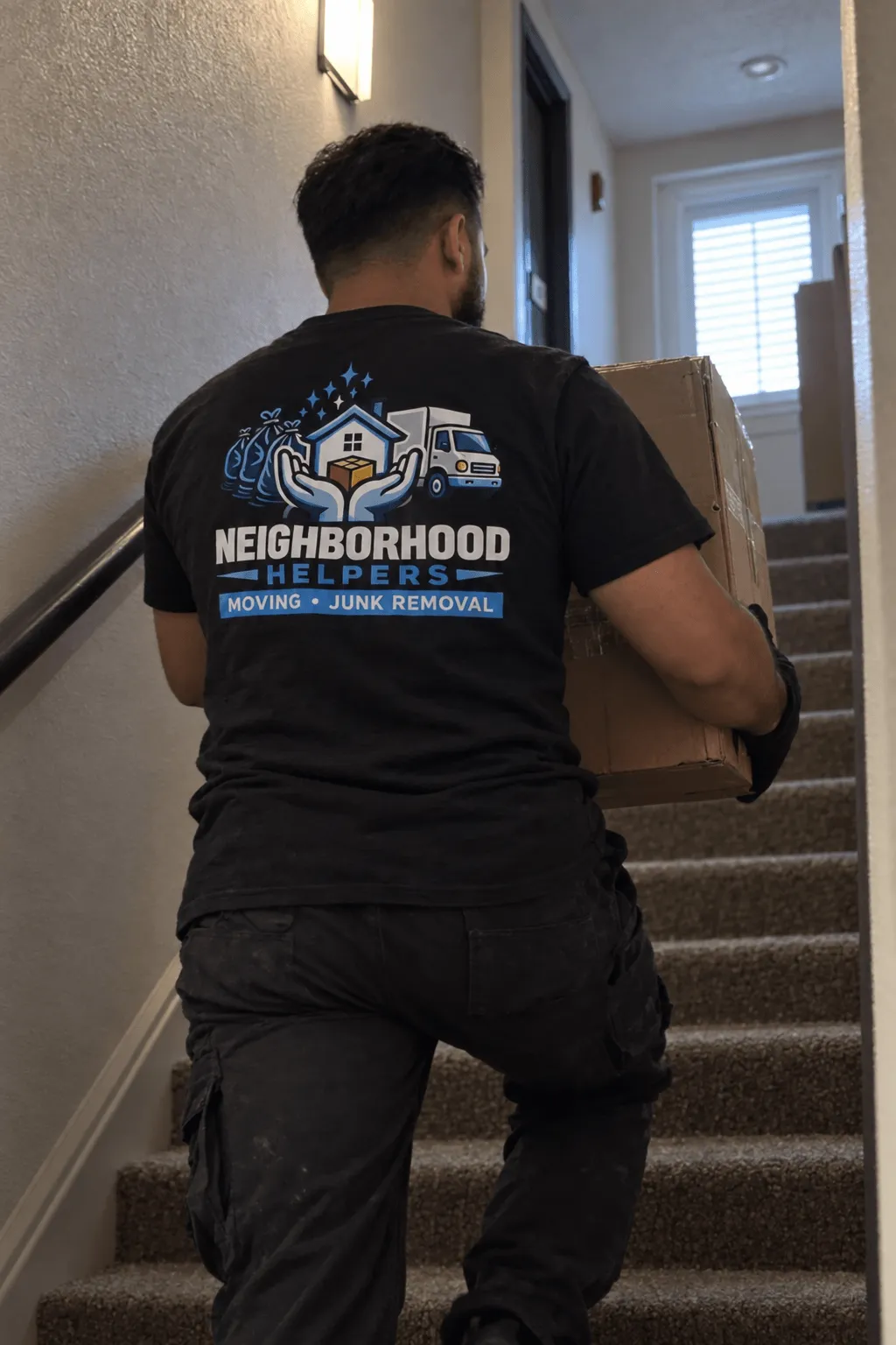 Worker wearing a black Neighborhood Helpers shirt carrying a cardboard box up carpeted stairs inside a residential building.