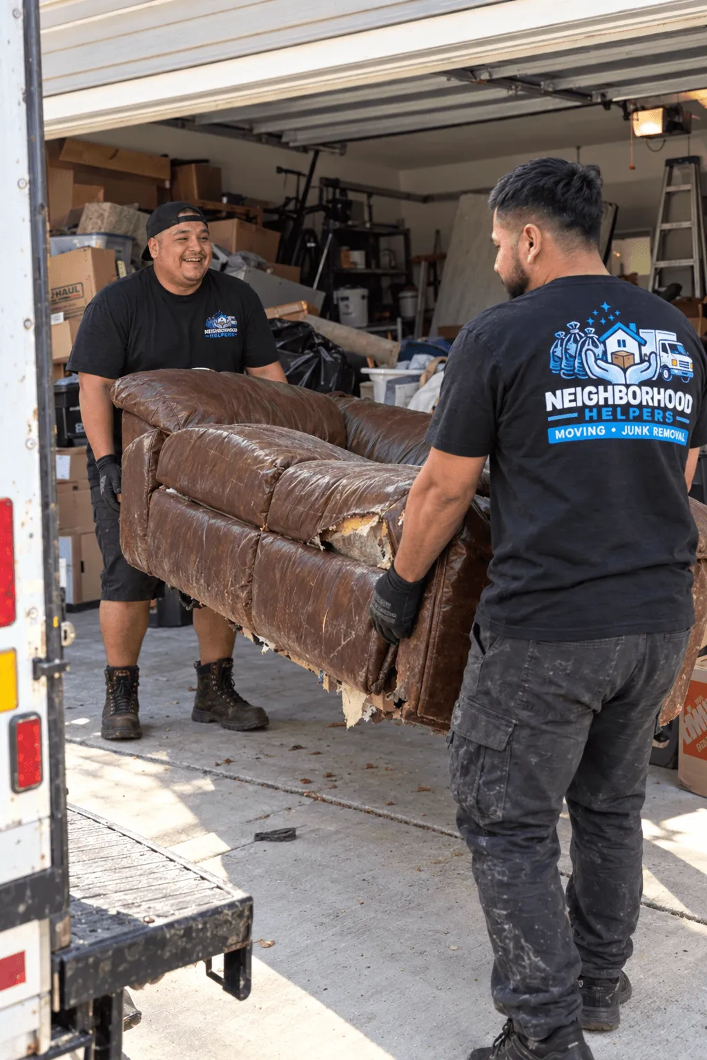 Two men wearing black Neighborhood Helpers t-shirts carry a damaged brown leather couch from a garage filled with boxes and other items toward a truck.