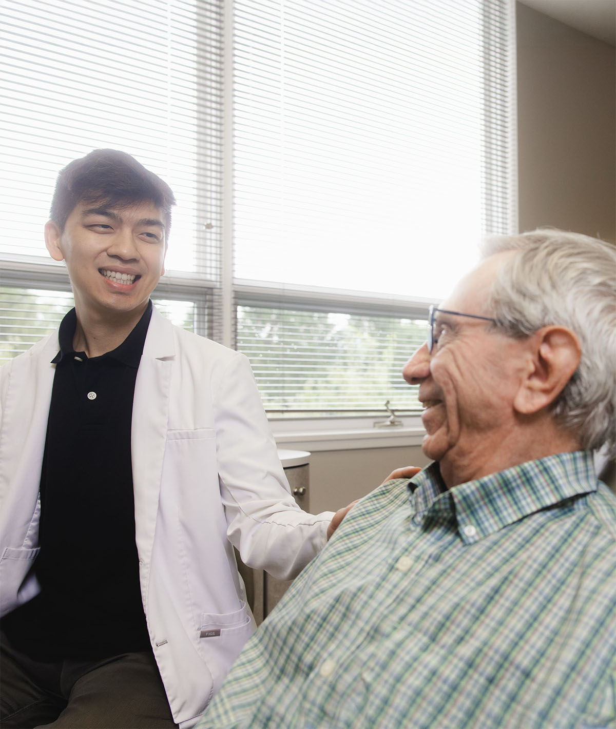 Young male doctor in a white coat smiling and talking with an elderly man wearing glasses and a green plaid shirt in a bright room with large windows.