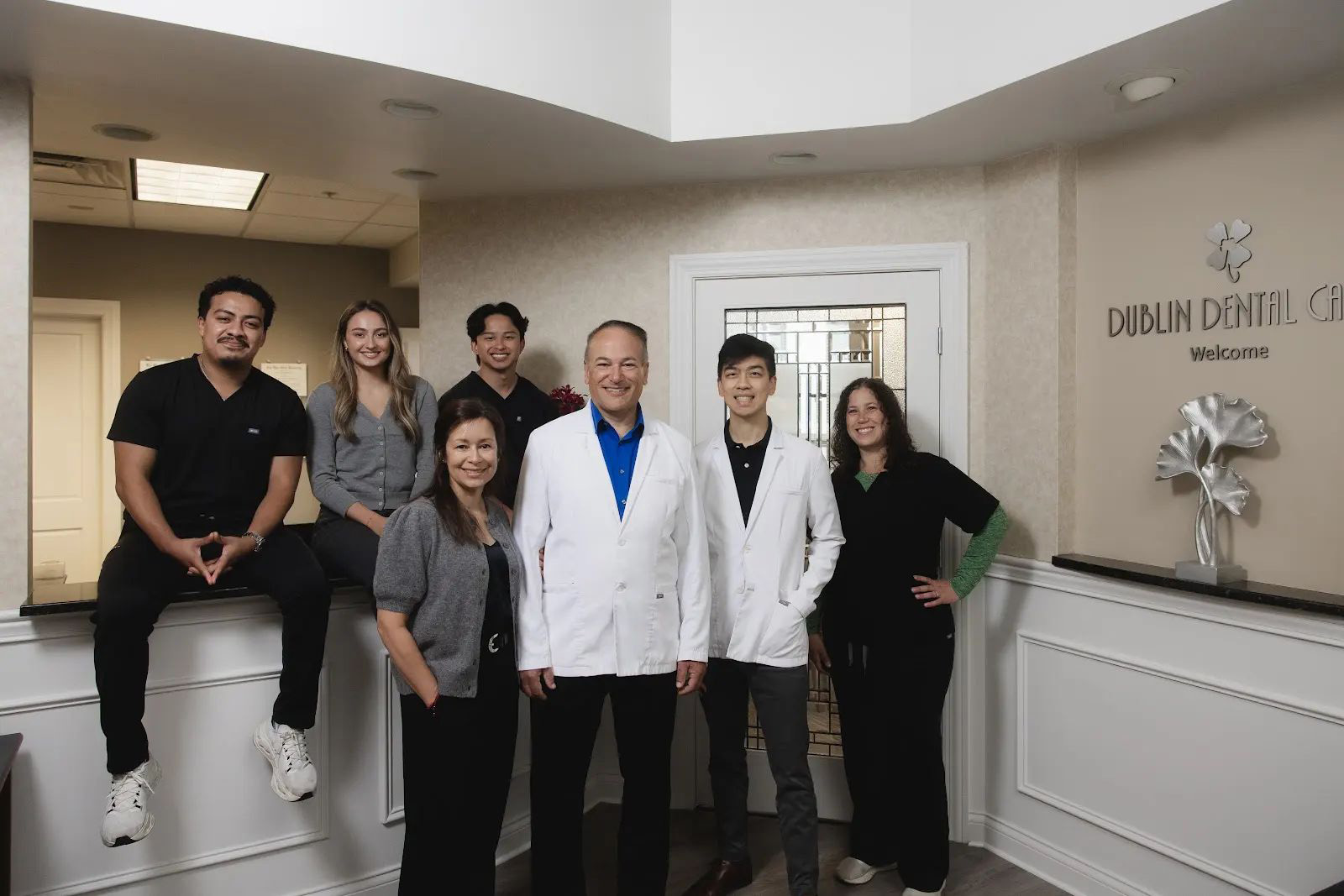 Group portrait of six dental professionals standing and one sitting on a counter in the reception area of Dublin Dental Care, smiling at the camera. The sign 'Dublin Dental Care Welcome' is visible on the wall with decorative silver flower sculptures.