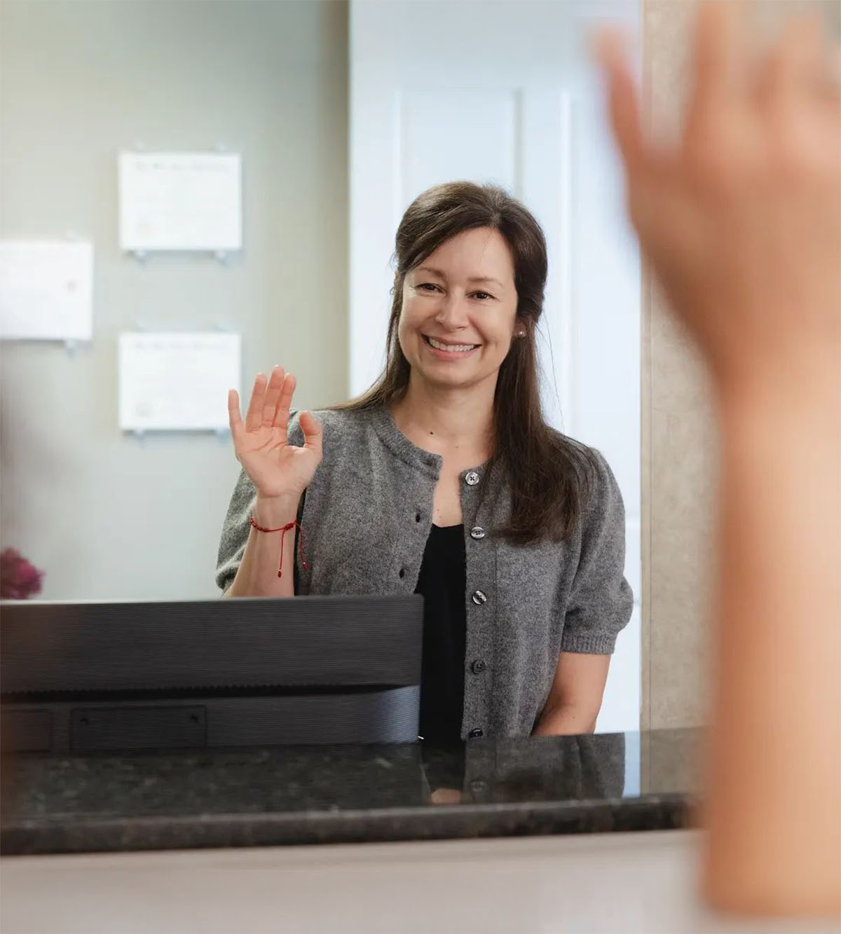 Smiling woman with long brown hair wearing a grey cardigan and black top, waving with her right hand behind a counter with a computer monitor.