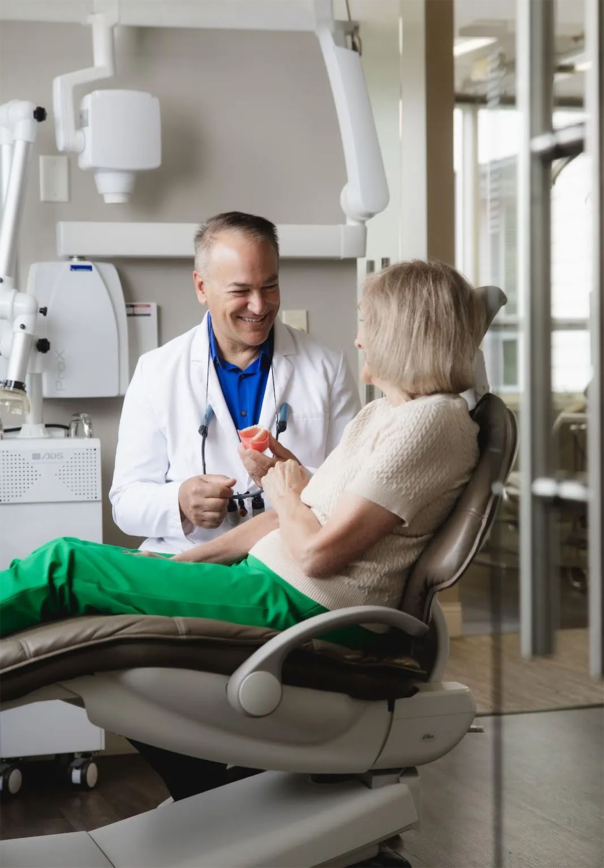 A male dentist in a white coat smiling and holding a dental mold while talking to a senior woman seated in a dental chair in a modern dental office.