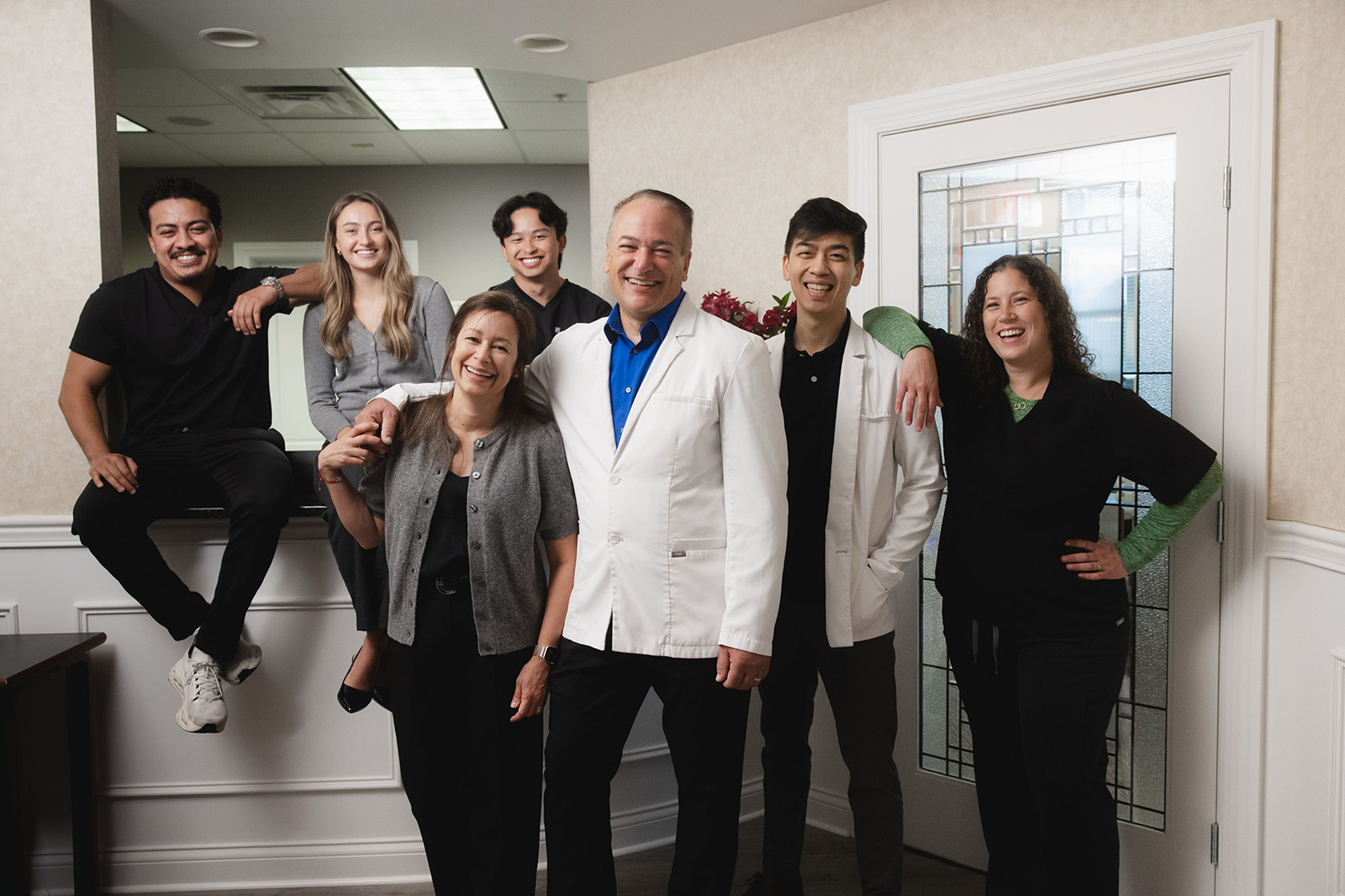 A group of seven diverse smiling medical professionals standing and sitting together in a well-lit office with beige walls and a decorative glass door.