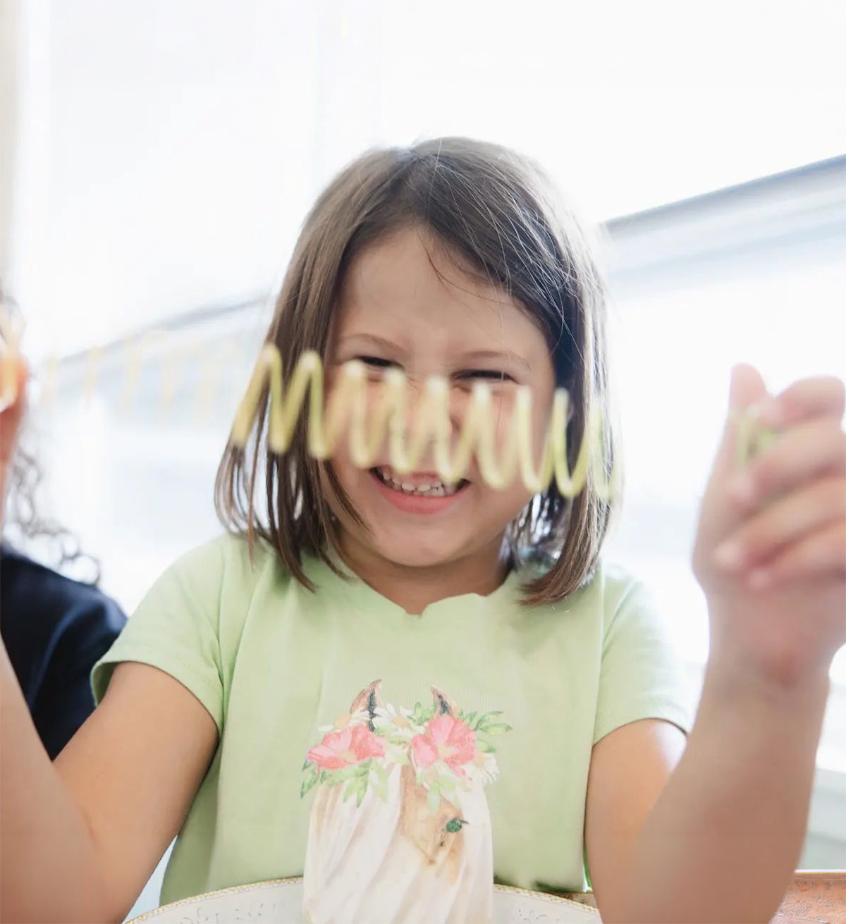 A smiling young girl with shoulder-length brown hair wearing a light green shirt with a horse and flower design, holding a yellow spring toy stretched in front of her face indoors near a window.