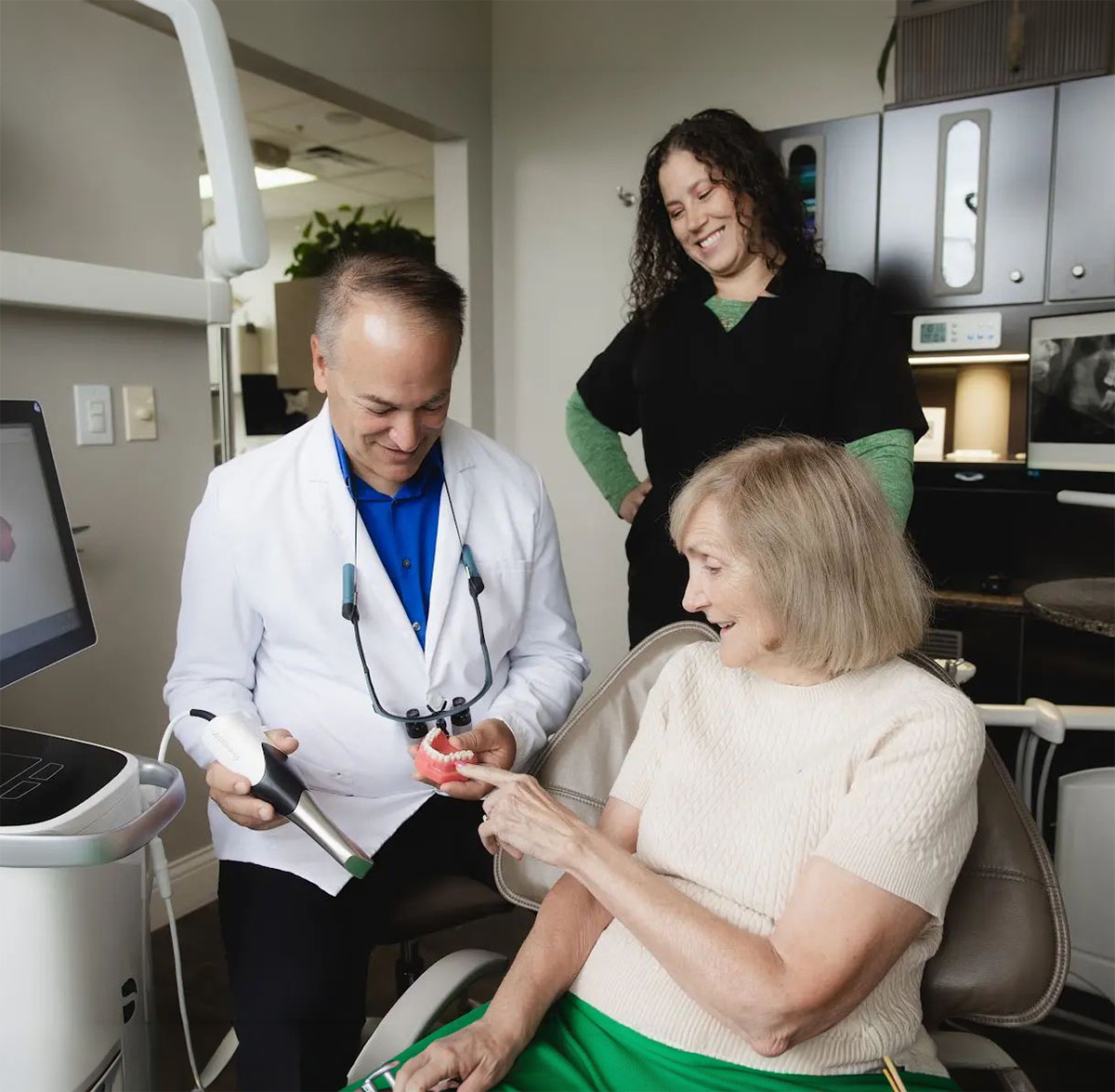 Dentist in a white coat holding a dental model and a scanning device while explaining to a seated elderly woman who is pointing at the model, with a dental assistant standing and smiling nearby in a dental office.