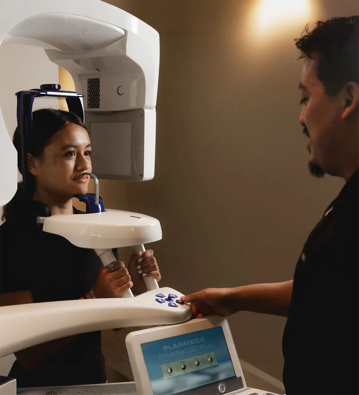 A woman undergoing a dental 3D imaging scan with her head positioned in a Planmeca ProMax 3D Plus machine while a technician operates the machine's control panel.