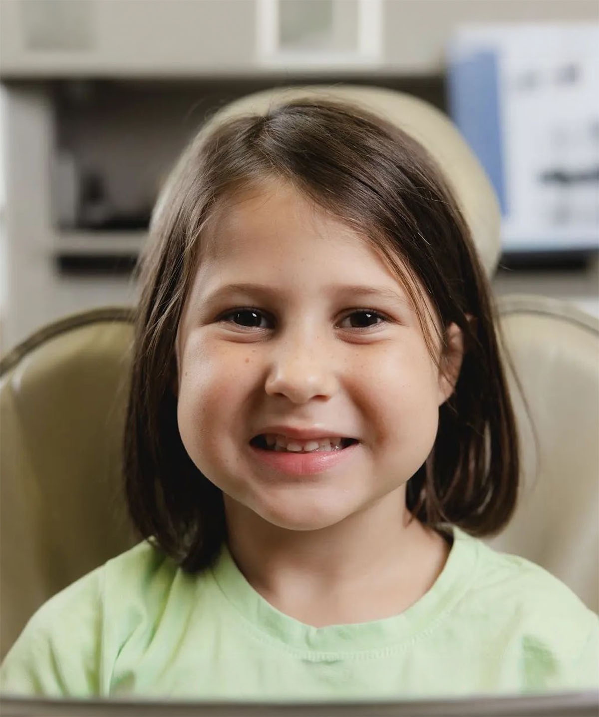 Smiling young girl with shoulder-length brown hair wearing a light green shirt, sitting on a beige chair indoors.
