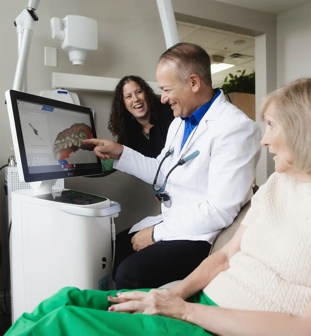A male dentist in a white coat is pointing at a 3D dental image on a screen while explaining to an elderly female patient seated beside him, with a woman standing behind them smiling.