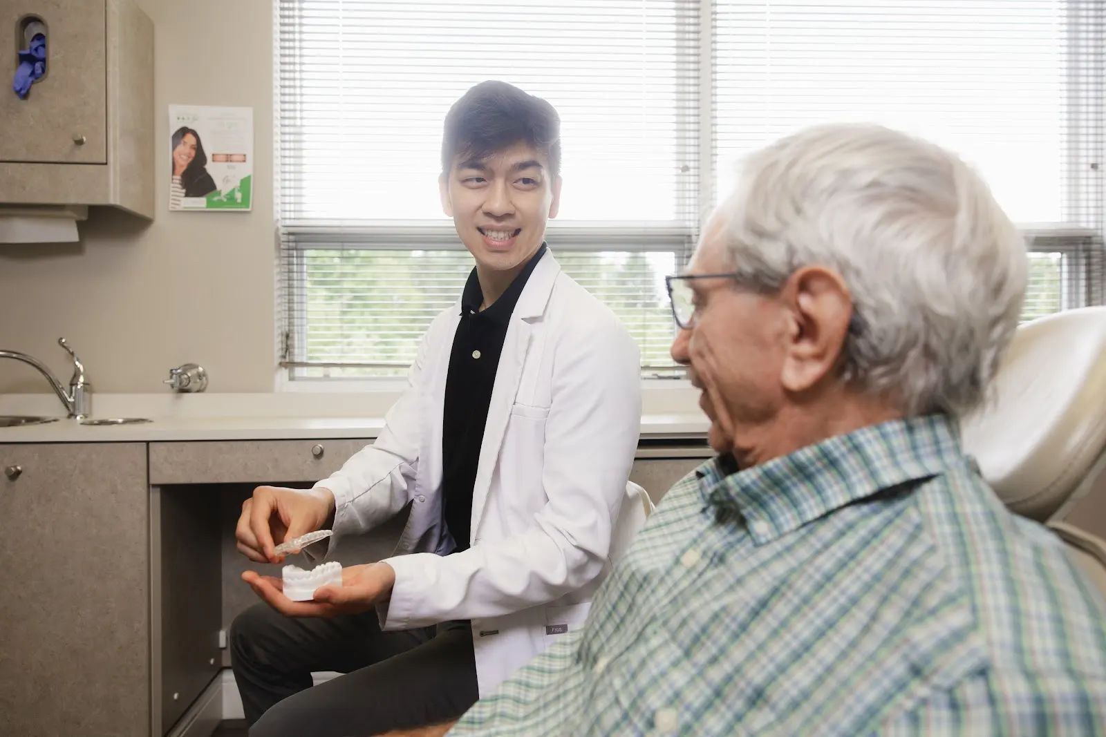 A young male dentist in a white coat shows a dental model with clear aligners to an elderly male patient sitting in a dental chair, in a well-lit dental office.