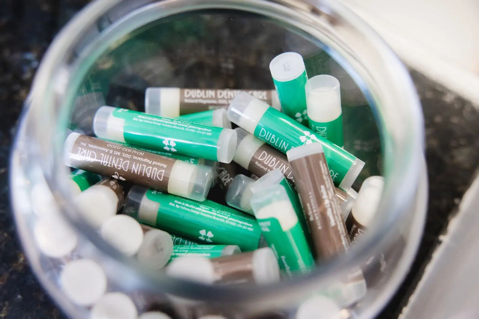 Glass jar filled with green and brown tubes of Dublin Dental Care lip balm on a dark surface.