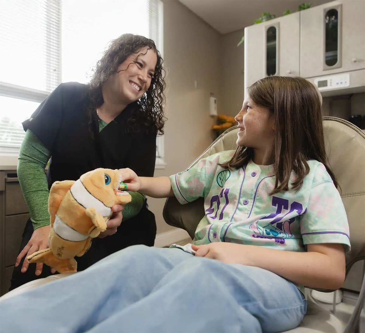 A smiling female dental professional wearing a black uniform and green undershirt interacts with a young girl seated in a dentist chair, holding a clownfish plush toy while the girl touches the toy's mouth.